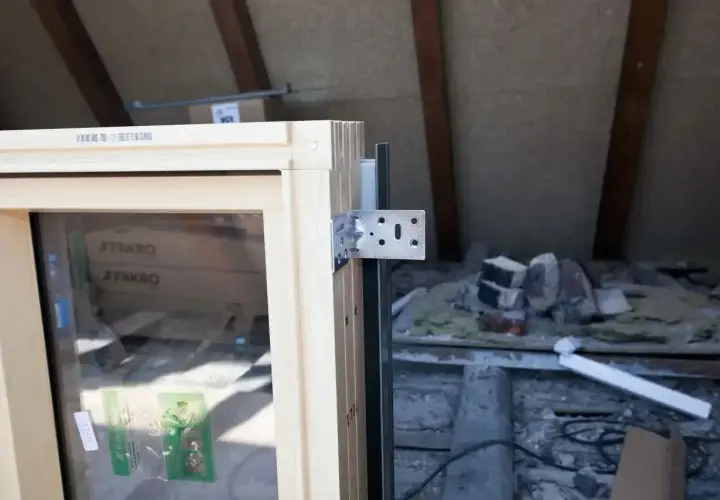 Drewniane okno dachowe z metalowym kątownikiem, gotowe do montażu. W tle widać materiały budowlane i elementy konstrukcji poddasza.