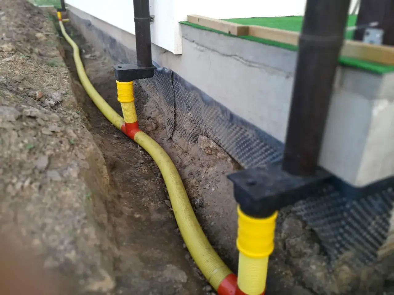 Instalacja odprowadzenia wody z rynien, ż&oacute;łty wąż w wykopie przy fundamentach. Przepisy dotyczące odprowadzenia wody z rynien są kluczowe dla ochrony budynku.
